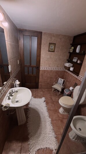 A bathroom with a white sink, a toilet, and a shower area. There is a wooden door, a framed picture on the wall, and shelves with toiletries. - CASA SANTA FE HOUSE (Luján de Cuyo)