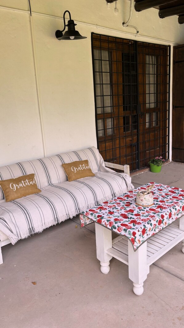 A white sofa with striped cushions and two pillows with the word "Gratitude" on them, a white table with a floral tablecloth, and a green pot with flowers on the porch. - CASA SANTA FE HOUSE (Luján de Cuyo)