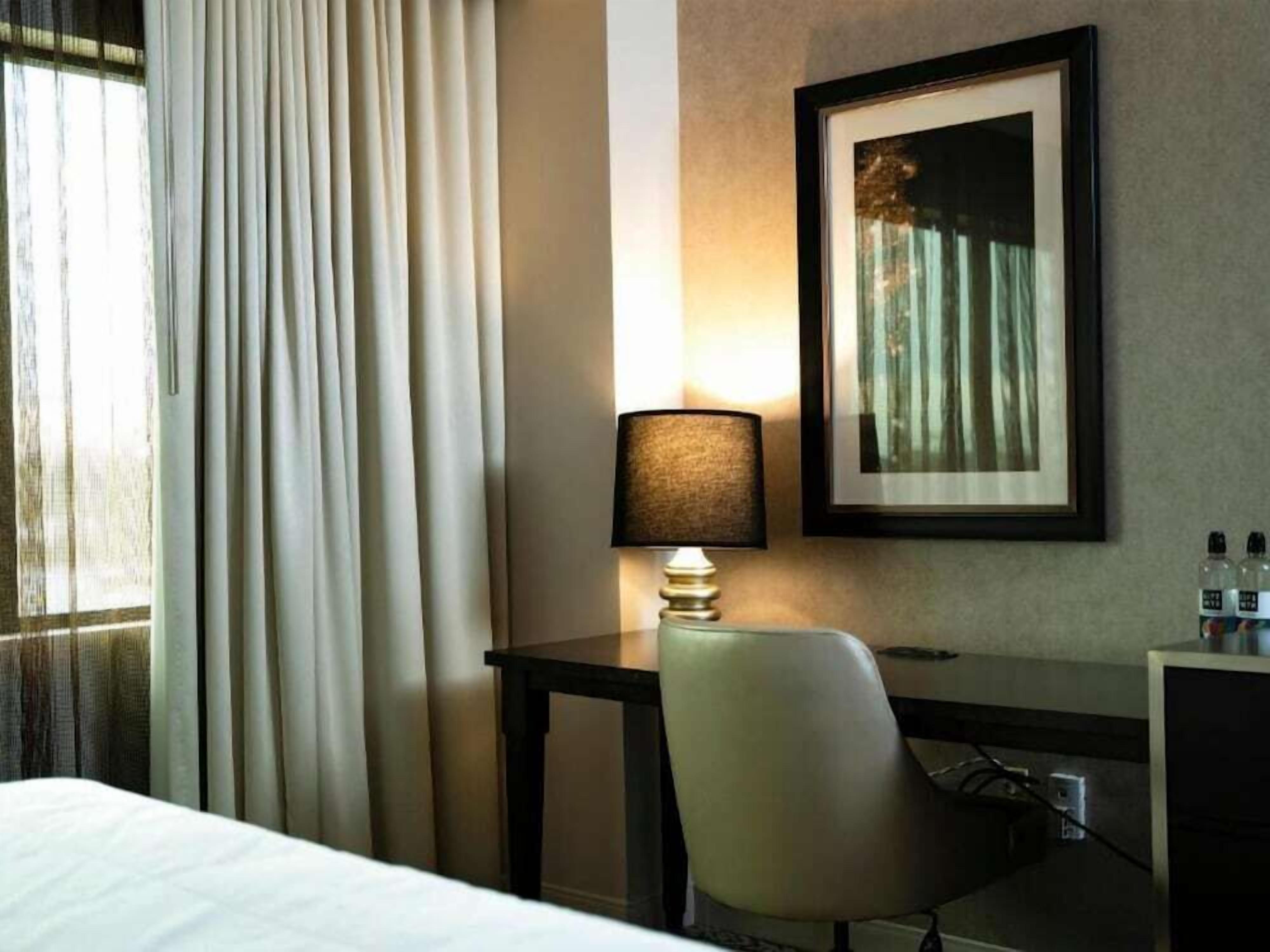Ein Hotelzimmer mit einem Bett, einem Schreibtisch mit Lampe, einem Sessel und einem gerahmten Bild an der Wand.