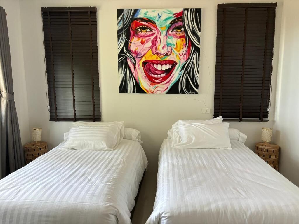 A room with two beds, a large painting on the wall, and a window with blinds.
