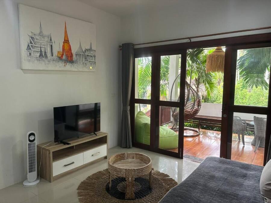 Una sala de estar moderna con televisor de pantalla plana, un mueble bajo para la TV de madera, una mesa de centro tejida y un gran ventanal corredizo que da a un patio exterior con una silla de mimbre y un colgante de luz.