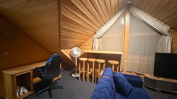 Un acogedor cuarto ático con techo inclinado de madera, un escritorio con una silla, una pequeña mesa con taburetes, un sofá y un televisor.