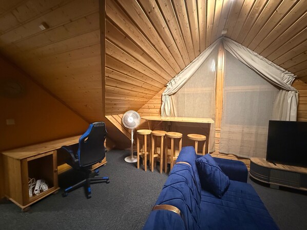 A cozy attic room with a wooden sloped ceiling, a desk with a chair, a small table with stools, a sofa, and a television.
