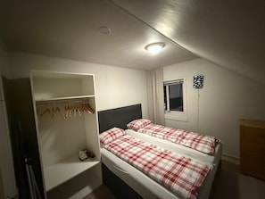 Um quarto pequeno e bem iluminado, com duas camas, um guarda-roupa e uma janela.