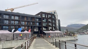 Um edifício moderno com vários andares, um cais que se projeta sobre a água e montanhas ao fundo.