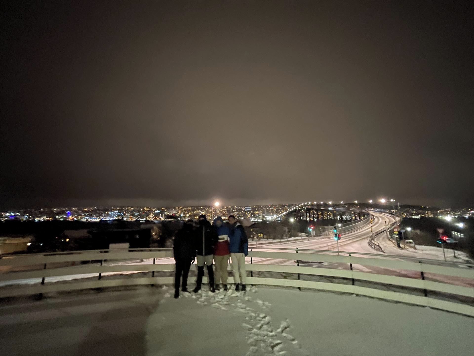 Yökuva kaupunkimaisemasta, otettu korkealta paikalta. Kuvassa näkyy lumipeitteinen alue, jossa on kaide, sekä liikennevaloin varustettu tie.