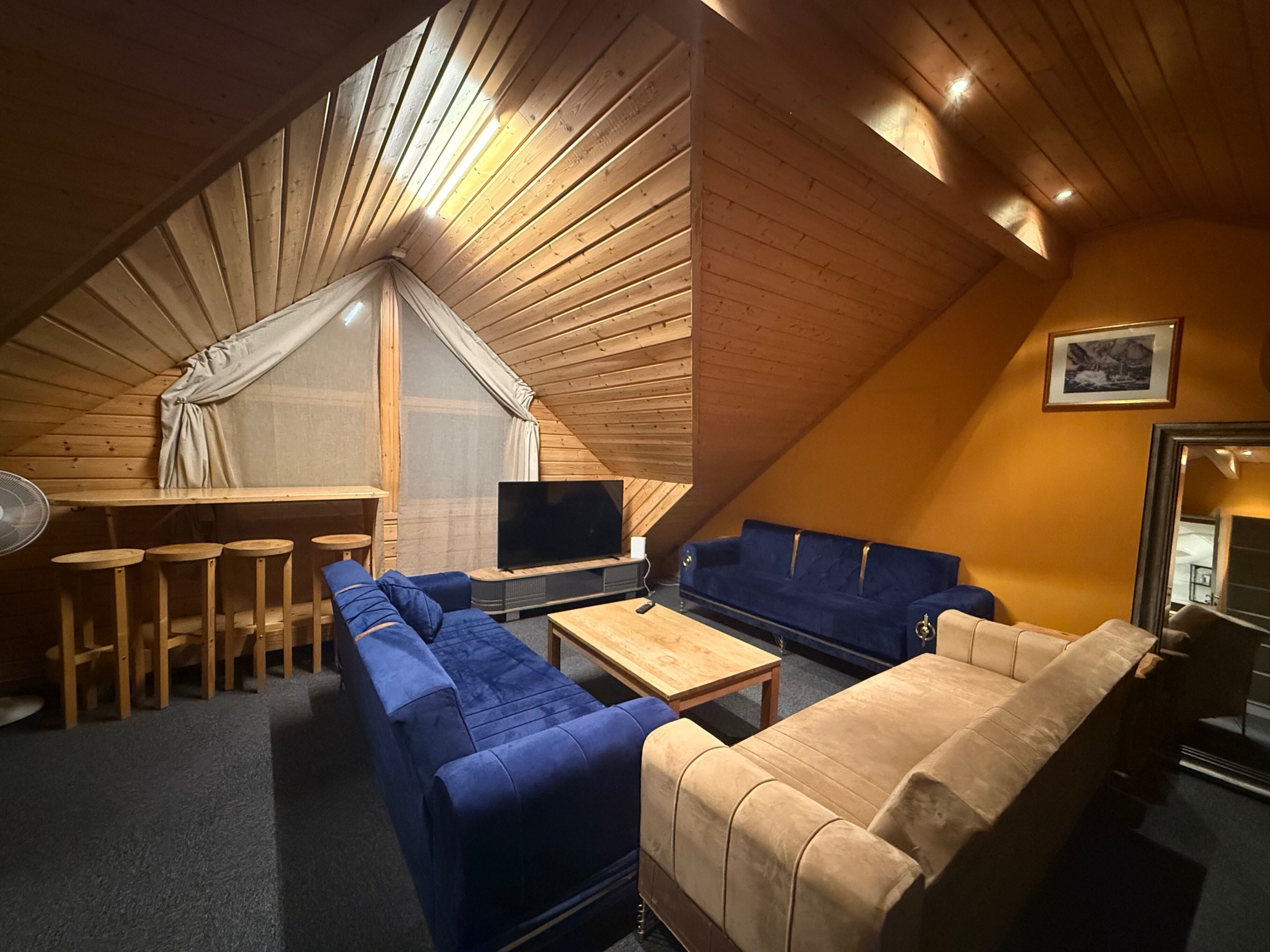 A cozy attic room with a wooden sloped ceiling, a large window with a white curtain, a TV, a blue sofa, a beige sofa, and a small wooden table.