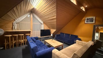 A cozy attic room with a wooden sloped ceiling, a large window with a white curtain, a TV, a blue sofa, a beige sofa, and a small wooden table.