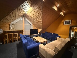 Um quarto de sótão aconchegante com teto inclinado de madeira, uma janela grande com cortina branca, uma TV, um sofá azul, um sofá bege e uma pequena mesa de madeira.