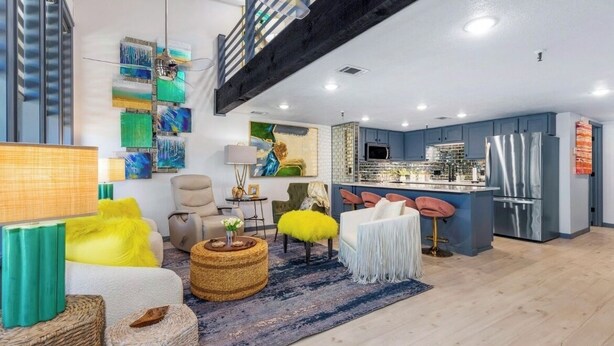 A modern living room with a sofa, armchair, and ottoman, a kitchen area with a bar, and a staircase with exposed beams.