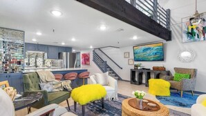 A spacious living area with a high ceiling, a staircase, and a kitchen in the background.