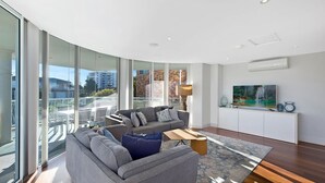 A modern living room with a grey sofa, a coffee table, and a TV on a white cabinet. Large windows offer a view of the city. - Skywaters Retreat 2 Bedroom (The Entrance)