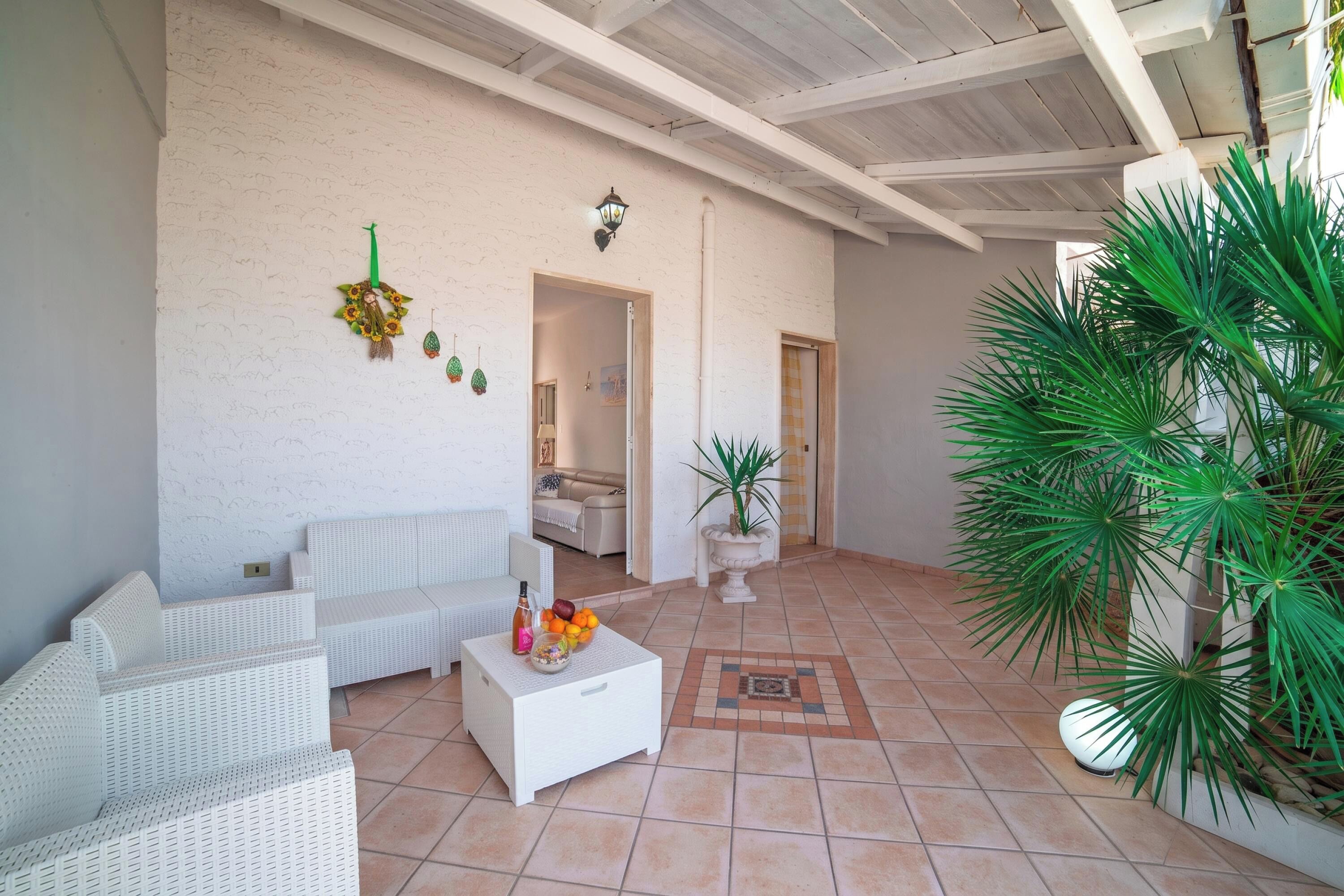 Ein geräumiger Patio-Bereich mit einem weißen Rattan-Sofa, einem weißen Couchtisch mit Früchten und einer Flasche, einer Topfpflanze und einem gefliesten Boden.