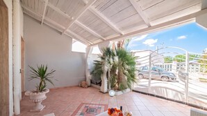 Un patio techado con suelo de baldosas, una zona de estar con sillas, una mesa con una botella y fruta, y vista a la calle a través de una reja de metal.