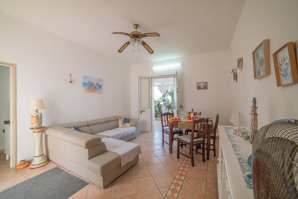 A living room with a sofa, a dining table with chairs, a ceiling fan, and a door leading to another room.