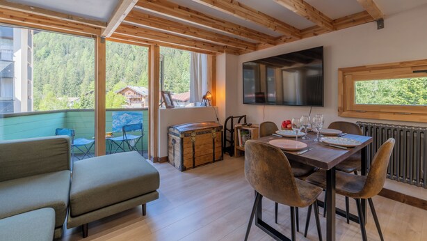 A spacious room with a wooden ceiling, a dining table set with plates and glasses, a sofa, and a large window overlooking a forested area.