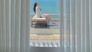 A person sitting on a wooden bench by a window with sheer curtains, overlooking a beach and the sea. - 3B MAISON Homestay & Villa (Mui Ne)