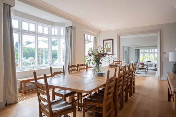 Un comedor amplio con una gran mesa y sillas de madera, ventanas grandes con cortinas y vista a áreas verdes.