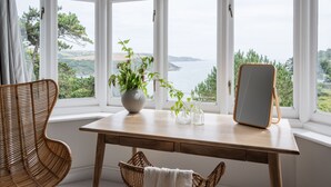A room with a wooden table, wicker chair, and a view of the sea through large windows. - The Wood -  a house that sleeps 15 guests  in 7 bedrooms (Salcombe)