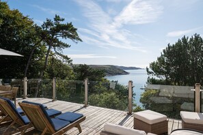 Terraza de madera con sillas de descanso y barandilla de vidrio con vista a un cuerpo de agua y colinas.