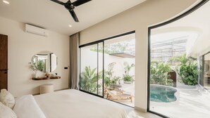 Un dormitorio moderno con una cama grande, ventilador de techo, espejo redondo y vista a un área de piscina exterior con plantas y mobiliario.