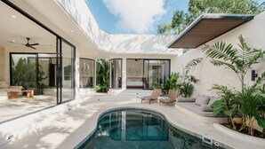 Un área de piscina moderna al aire libre con un salón con paredes de vidrio, un jacuzzi y una zona de estar con cojines y plantas.