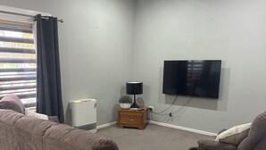 A living room with a beige carpet, two brown sofas, a small wooden side table, a floor lamp, and a flat-screen TV mounted on the wall. - Awesome Central Horsham Home (Horsham)
