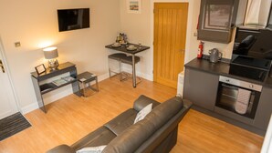 A compact living space with a sofa, a small dining table with chairs, a kitchenette, a TV mounted on the wall, and a wooden door. - Tigh Na Cille Studio (Taynuilt)