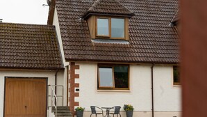 A two-story house with a tiled roof, a wooden door, and a small patio with two chairs and potted plants. - Tigh Na Cille Studio (Taynuilt)