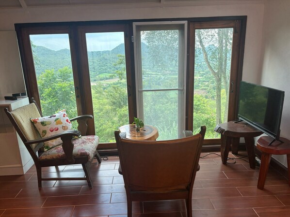 A room with a large window, a television, a chair, a small table with a plant, and a wooden floor. - Tawan Ja Resort (Pak Chong)