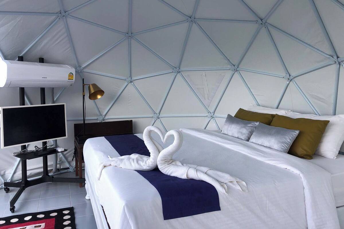 A modern hotel room with a geometric ceiling design, a bed with white linens and decorative swan-shaped towels, a bedside table with a lamp, and a flat-screen TV.
