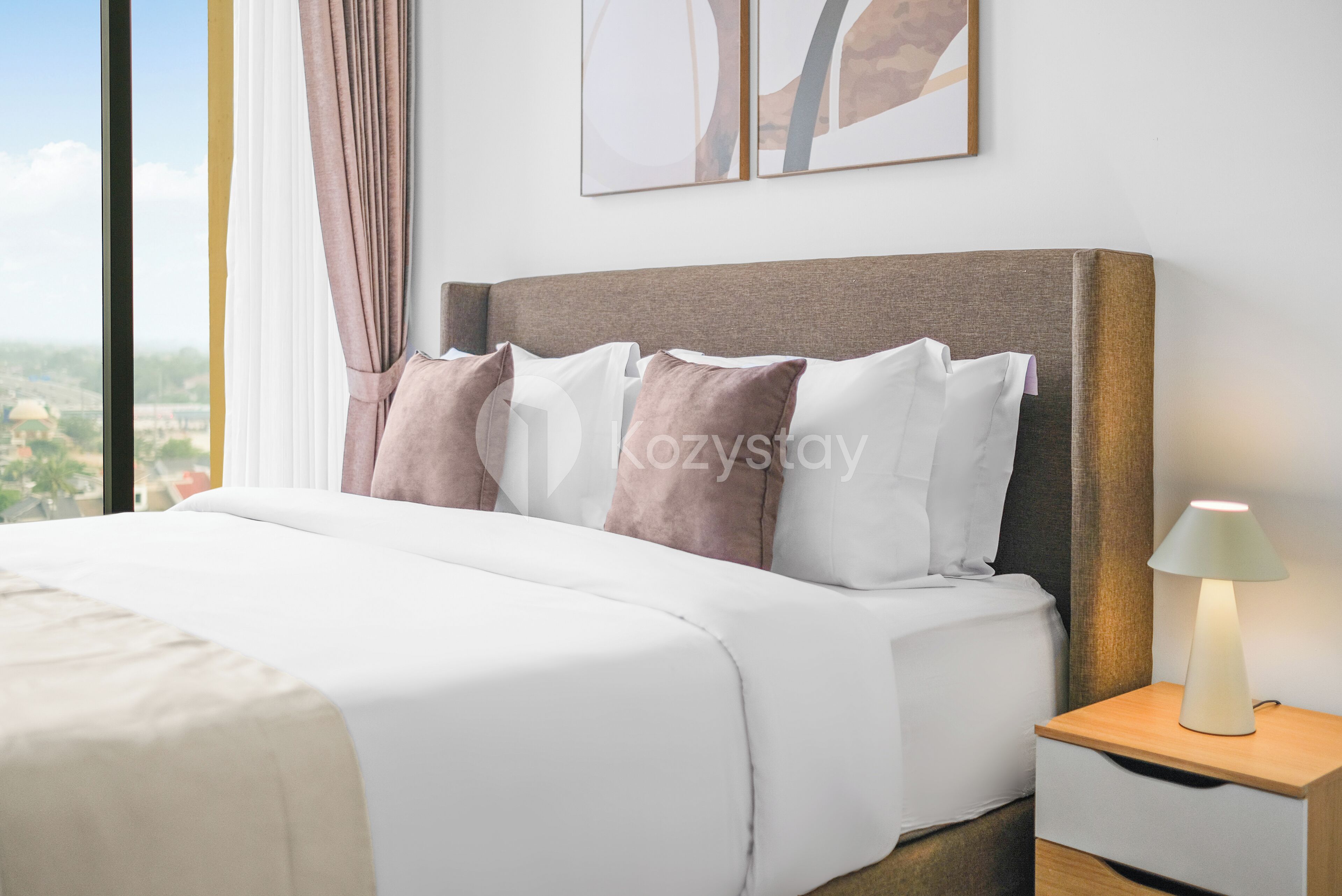 Una cama bien hecha con ropa de cama blanca y beige, una mesita de noche con lámpara, y vista a la ciudad a través de una ventana.