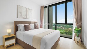 Een moderne slaapkamer met een groot bed, een nachtkastje met een lamp, uitzicht op groen en gebouwen door het raam, en een plant in een pot op de vensterbank.