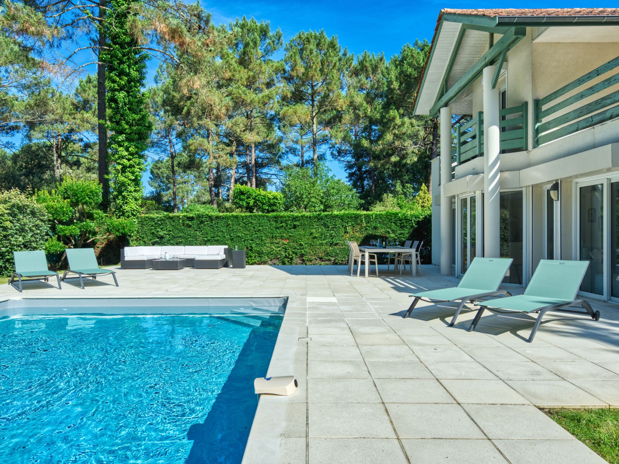 A pool area with lounge chairs and a dining table, surrounded by a hedge and trees.