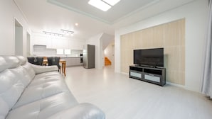 Un salon moderne avec un canapé en cuir blanc, un téléviseur à écran plat posé sur un meuble noir, et une cuisine en arrière-plan.