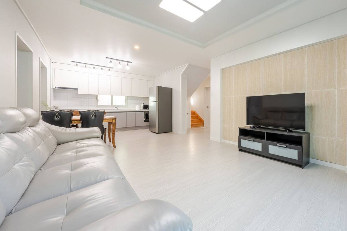 Uma sala de estar moderna com um sofá de couro branco, uma TV de tela plana em um suporte preto, e uma cozinha ao fundo.