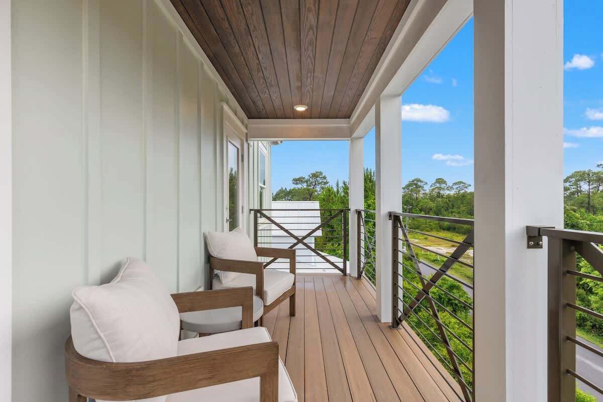 A covered porch with wooden flooring, white walls, and a metal railing. There are two white cushioned chairs and a small table. The porch overlooks a green landscape with trees and a road.