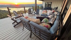 Una terraza con sofá, sillas y una mesa, con vistas a una masa de agua y colinas al atardecer.