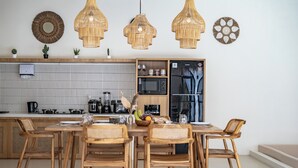 Una cocina moderna con armarios de madera, un microondas integrado y un refrigerador. Hay lámparas colgantes de mimbre, un espejo redondo y arte decorativo en la pared.