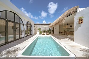 Zona de piscina con una piscina azul cristalina, un techo de paja y un edificio moderno de color blanco con grandes puertas de vidrio.