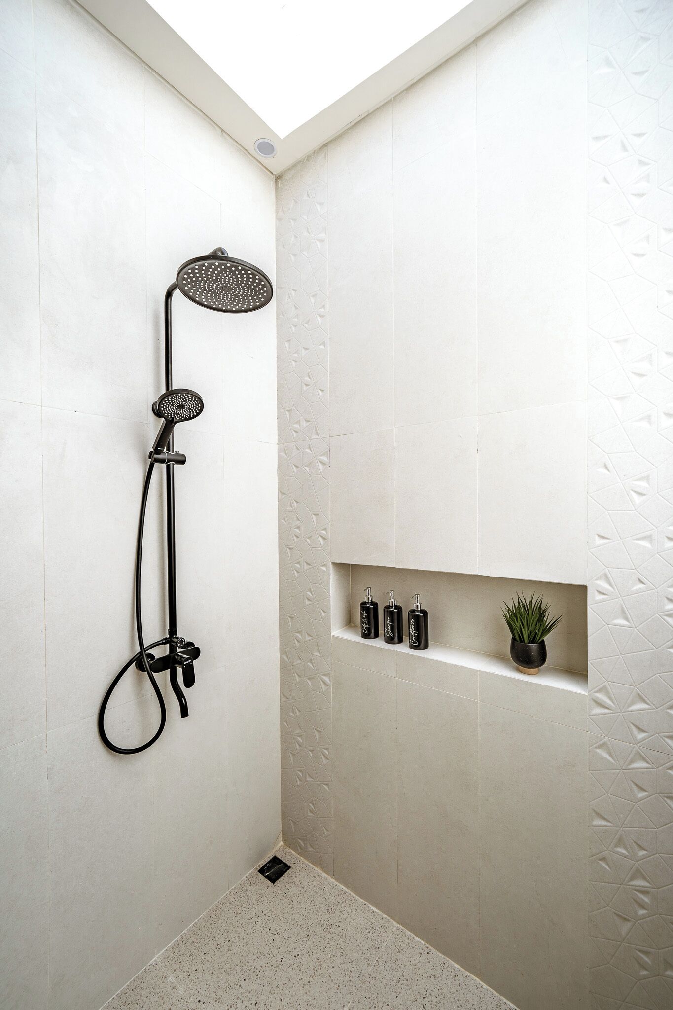A shower area with a handheld showerhead, a wall-mounted shelf with toiletries, and a small potted plant.