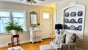 Una sala de estar con un sillón estampado, una mesita, una planta en maceta, un aparador y una colección de platos decorativos en la pared.