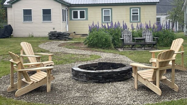 Una casa con fogón en el jardín, dos sillas Adirondack y una pila de leña.