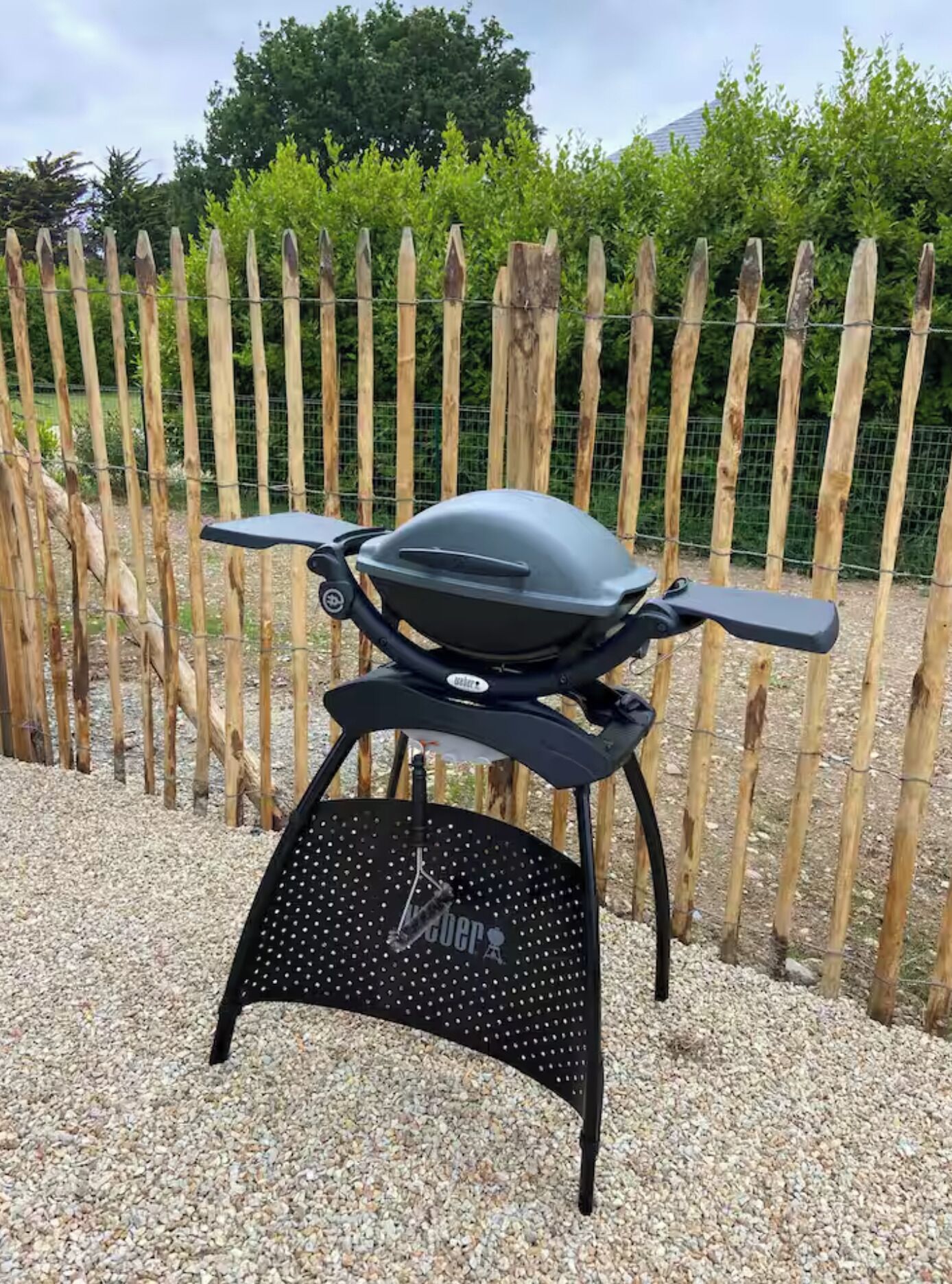 A portable grill with a black body and perforated base, set on a gravel surface with a bamboo fence and greenery in the background.
