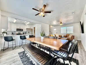 A modern kitchen with white cabinets, a dining area with a wooden table and black chairs, and a ceiling fan. - Glendale Retreat | Hot Tub Near Zion & Bryce (Glendale)