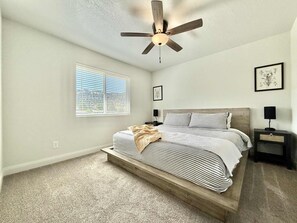Une chambre à coucher comprenant un lit, un ventilateur de plafond, une table de chevet, une fenêtre avec des stores et un tableau encadré accroché au mur.