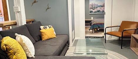 Un salon moderne avec un canapé gris, une table basse en verre et un fauteuil en cuir marron. Un tableau est accroché au mur et une porte donne accès à une autre pièce.