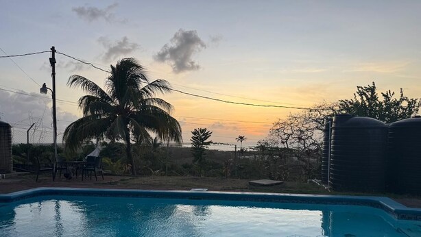 A swimming pool with a sunset in the background, surrounded by palm trees and a fence. - Cozy 1-bedroom apartment in adorable Tobago with AC, WiFi (Tobago)