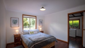 A bedroom with a wooden bed, two bedside tables with lamps, framed botanical prints on the wall, and a view of the outdoors through a window.