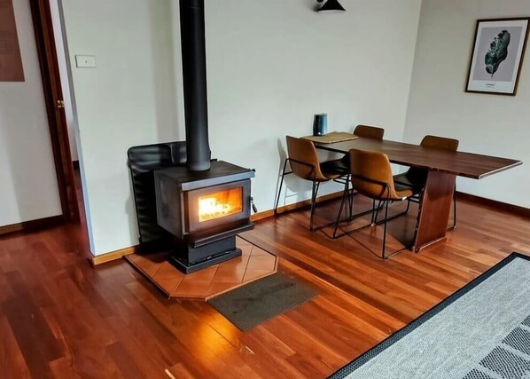 A room with a wood-burning stove, a dining table with chairs, and a rug on the floor.